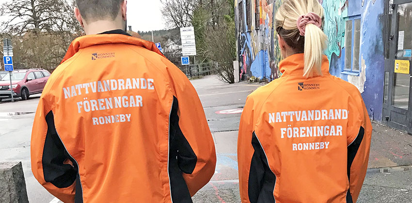 Två personer går på en gata. De har jackor med texten Nattvandrande föreningar Ronneby på baksidan.