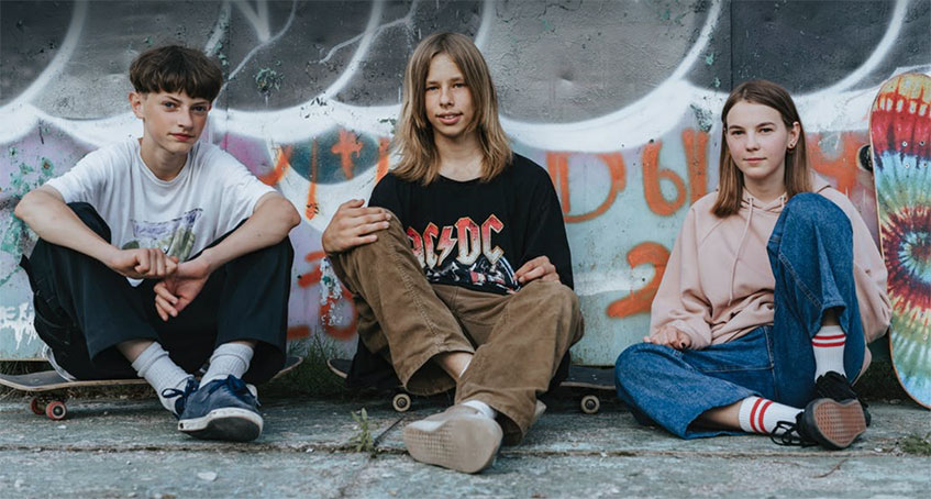 Tre ungdomar sitter utomhus på marken med skateboard. Bakom finns en vägg med graffitimålning.