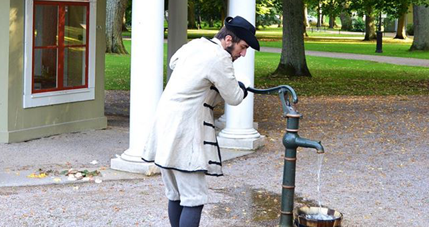 En man i tidsenliga kläder från den forna brunnsepoken pumpar upp vatten ur Brunnskällan i Brunnsparken i Ronneby.