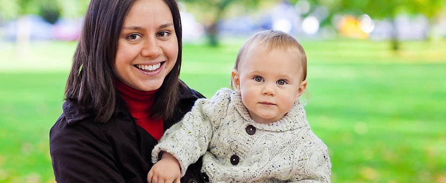 En ung mamma med ett barn i famnen, ca 7 månader, bilden tagen med grönska bakom, båda tittar glatt in i kameran