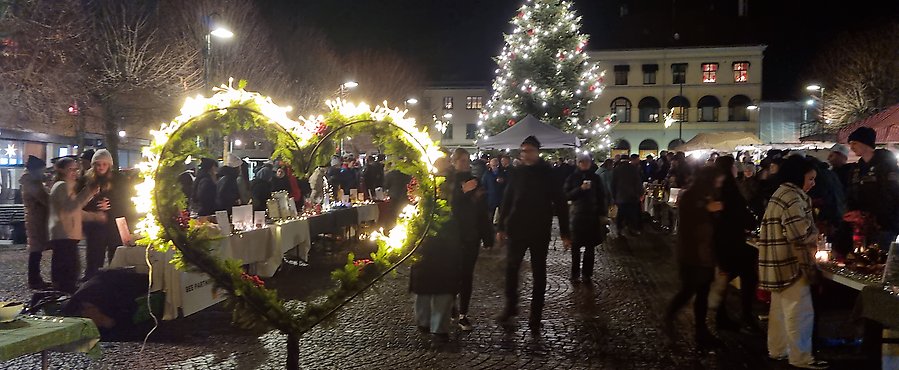 Julaktivitet på torget en kväll, torgets julgran i bakgrunden, ett hjärta i förgrunden, många människor i mörka siluetter
