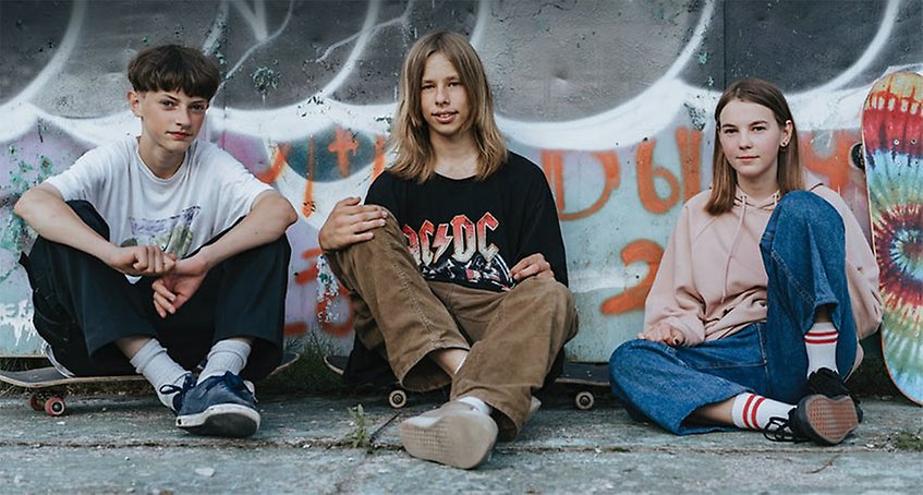 Tre ungdomar sitter utomhus på marken med skateboard. Bakom finns en vägg med graffitimålning.
