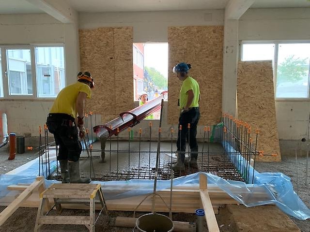 Två byggarbetare gjuter en betongplatta inomhus. Armeringsjärnet syns delvis. En tredje byggarbetare syns genom ett fönster