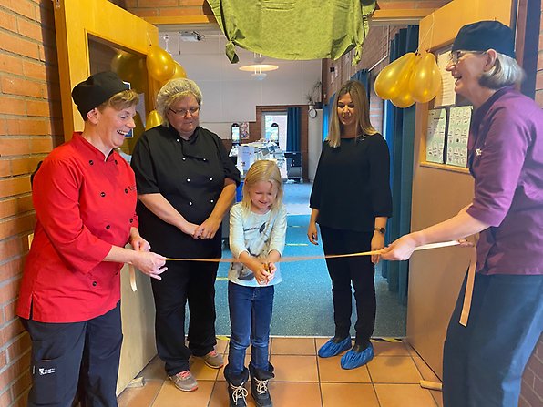 Fyra personer och ett barn. Barnet håller i en sax och ska strax klippa invigningsbandet till den nya matsalen.
